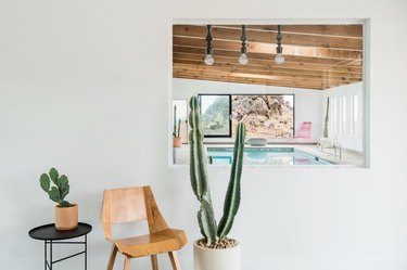 An interior window looking into pool area with wooden chair, side table and potted cacti.