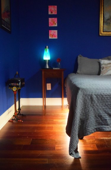 A bedroom corner with a lights emanating from under a bed on a hardwood floor. There is art on the walls, a small cast iron stand, and a wooden nightstand with a lava lamp on top.