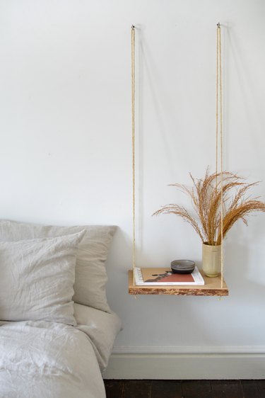 Wooden nightstand shelf with a vase of grasses hanging from two wall hooks next to a neutral bed and dark floor.