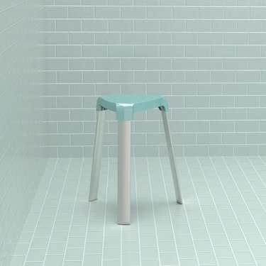 A three-legged alumnium stool with a light blue seat and grey legs sits in a corner with rectangular light blue tile.