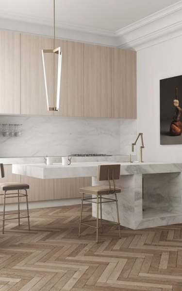 Chevron or zigzag patterned wood in a modern kitchen with marble island, pendant light, marble backsplash and blond wood cabinets.