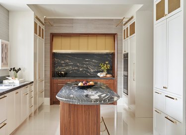 Oval kitchen island with marble counter, wooden base, white cabinets, glossy white flooring.