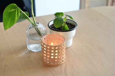 Cane candle holder on wooden table next to plant pot and glass vase.