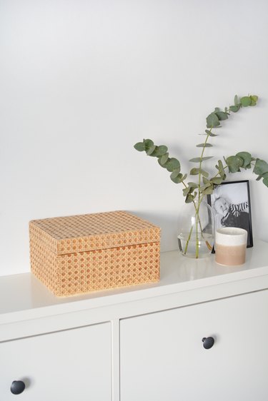 Wooden box covered in cabe on white unit with candle, framed photo and vase with greenery in background.