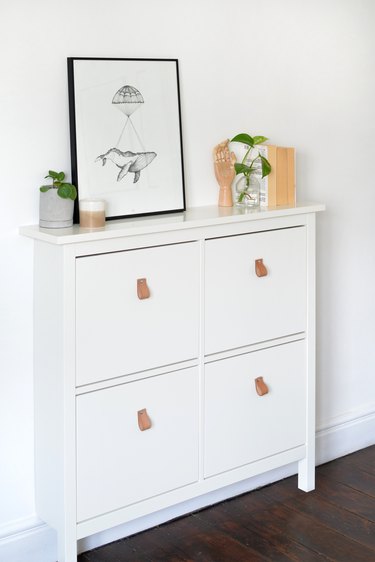 White IKEA shoe storage cabinet with framed poster, books and decorative items.