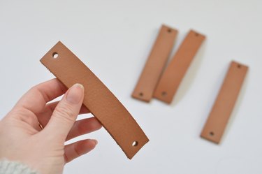 Hand holding piece of tan leather with a hole punched at each end.