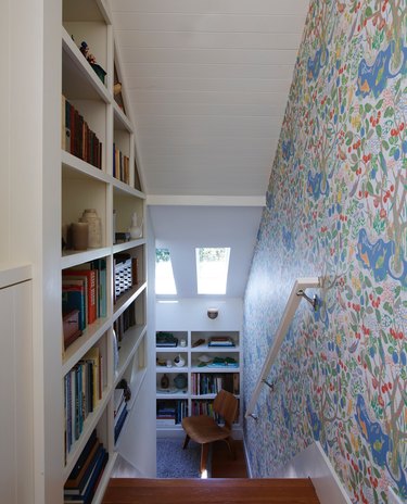 Staircase with bookcase and floral wallpaper down to room with skylights