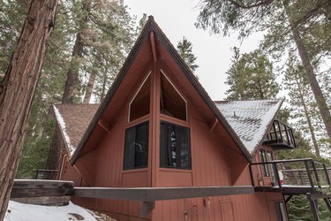A brown Midcentury cabin in the woods. The roof is multi v-shaped with snow. Four windows meet at an angle. Trees surround the home.