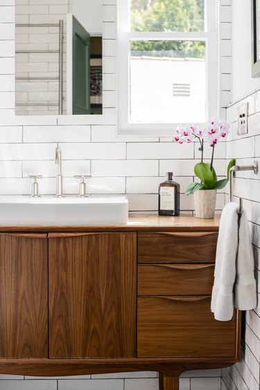 wood vanity with three drawers, white ceramic vessel sink with silver fixtures, rectangular mirror, window, white towel hung up, vase with orchids