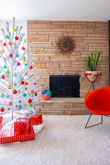 Christmas Tree Themes with Silver artificial Christmas tree, colored ball ornaments, red lounge chair, stone fireplace, white area rug, wrapped presents.