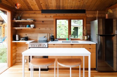Rustic kitchen with wood paneled walls, wood cabinets, white table, stainless steel electric stove and fridge, wood floors, open shelves.
