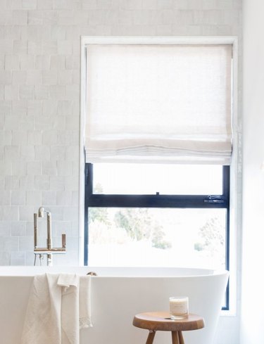 bathroom tub backsplash idea with zellige tile behind freestanding tub and window with Roman shade