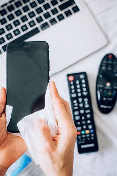 person cleaning iPhone with wipe and showing remotes and laptop on the counter