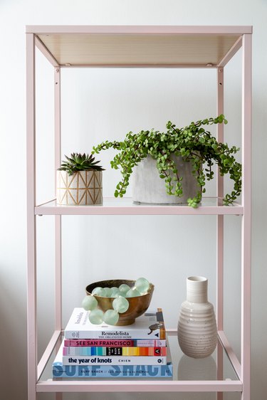 Pink metal shelf with books and plants and a white vase.