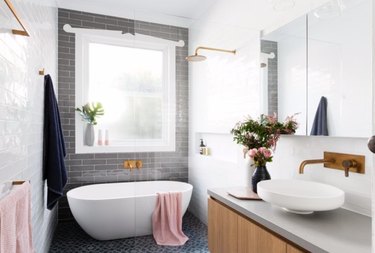 A white tub within a modern shower with grey tiles and brass shower head.