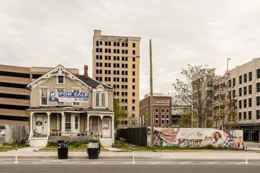 house with for sale sign near abandoned lot and high-rise buildings