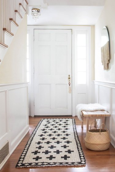 Painted jute rug in entryway hall next to bench, basket, and fringed mirror on wall.