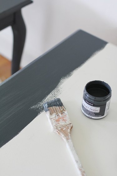 Paint brush and small can of slate colored paint on white furniture.