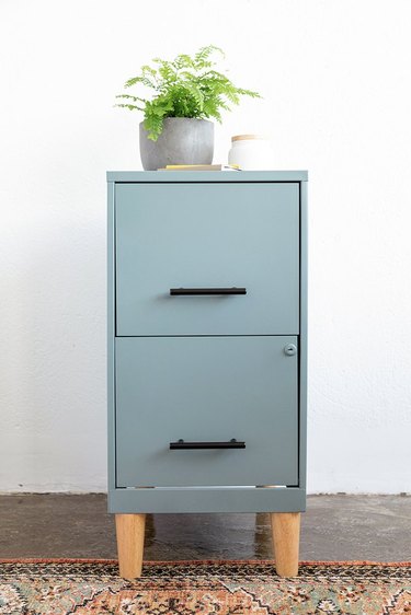 Teal colored filing cabinet with wood legs on rug with plant on top.