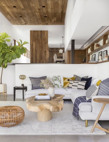 coastal living room with wood ceiling, concrete floors, tree trunk coffee table, white sectional with multi colored throw cushions, cane pouf, fiddle leaf fig plant.