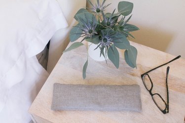 Gray linen eye pillow on top of nightstand with eyeglasses and flowers