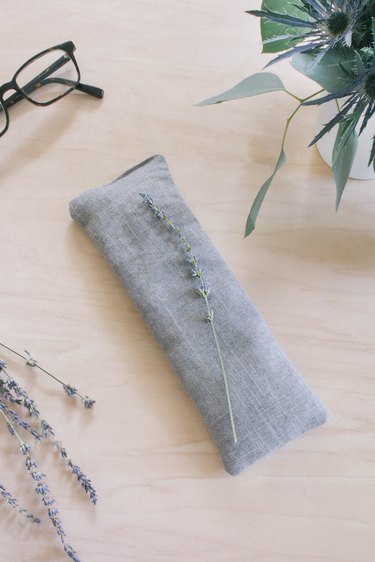 Gray linen eye pillow on top of nightstand with eyeglasses and flowers