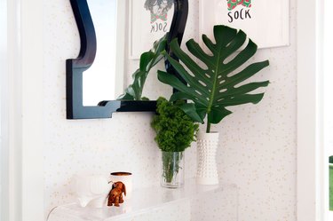 Minimalist entryway design with decorative mirror above clear console table with plant leaves in vases.