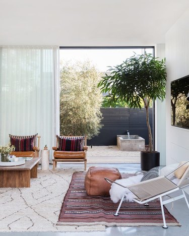 living room rug idea with big sliding glass doors in the background, in the foreground a wire chair and leather pouf sit atop a kilim rug layered over a flokati rug