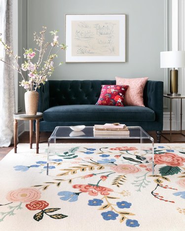 living room rug idea with a light pink rug with a large floral design, an acrylic coffee table sits on top and a dark velvet couch is in the background