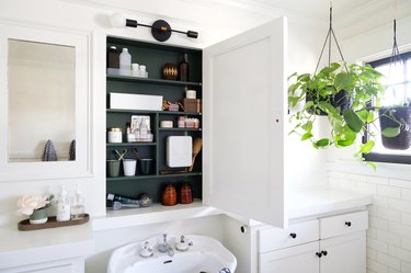 white bathroom, white subway tile, window with black trim, hanging plant, recessed medicine cabinet with painted wood green interior, white pedestal sink