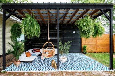 Black wood and metal modern pergola with sectional, hanging chair, plants and large outdoor rug.