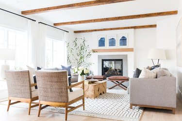 coastal living room with beige couch, wicker side chairs, tree trunk coffee table, wood beamed ceiling, fireplace, plant, white walls.