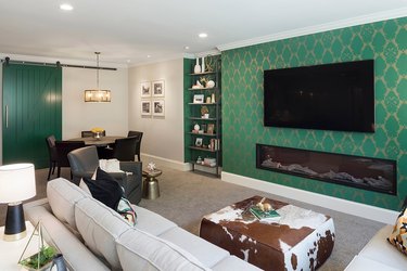 transitional family room with green flocked wall paper, flat screen tv, cowhide pouf, off-white couch.