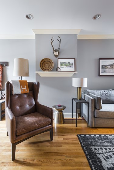 transitional family room with high back leather chair, gray couch, wood floors, side table, lamp, shelf with art and basket, antlers on wall.