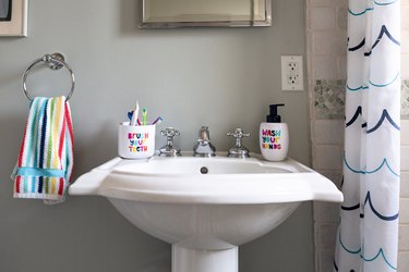 white pedestal sink, rainbow striped hand towel hung up, a toothbrush holder that says brush your teeth, a soap dispenser that says wash your hands
