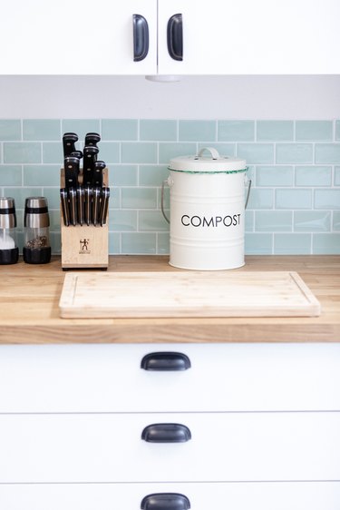 close up of kitchen wood butcherblock countertops with compost bin sitting on top and blue tile backsplash