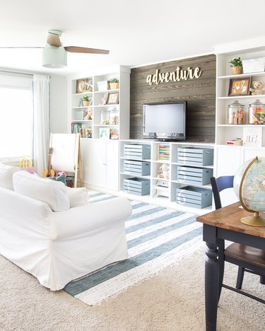 family room storage with white built in shelving with flat screen tv, storage bins, white couch, blue striped rug.