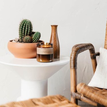 Cactus in terra cotta pot, P. F. Candle Company candle, ceramic vase, white end table, cane side chair, white pillow.