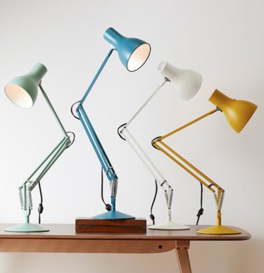 Four desk lamps in green, blue, white and yellow on top of wood table.