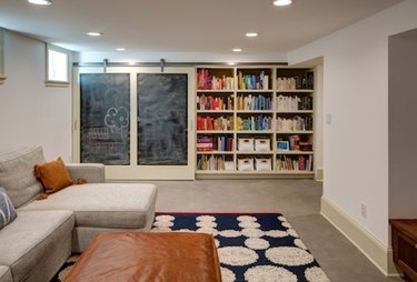 Recessed basement lighting in off white basement with beige sectional, leather ottoman, white and blue graphic area rug, built in bookcases and blackboard.