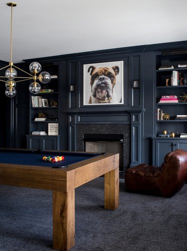 basement game room ideas with dark gray pool room, modern pool table, brass modern chandelier, leather lounge chair, built in book shelves, fireplace.