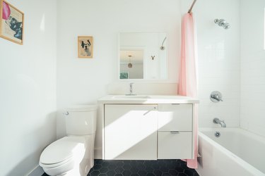 A white bathroom with a toilet, floating vanity, and a white bathtub/shower with a pink shower curtain