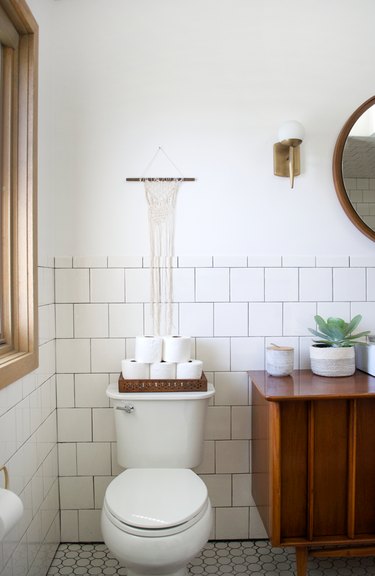 guest bathroom idea with toilet paper stacked in a try on the back of a toilet in a white-tiled bathroom