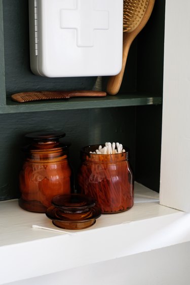 recessed medicine cabinet with painted green wood interior, glass jars of cotton balls, q-tips, white first aid kit, wooden brushes