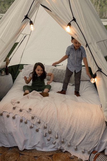 Two kids jumping on bed inside glamping tent