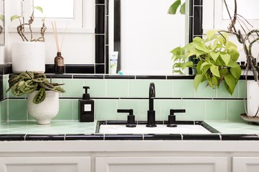 bathroom vanity with white cabinets, green and black ceramic tile, white planters with greenery