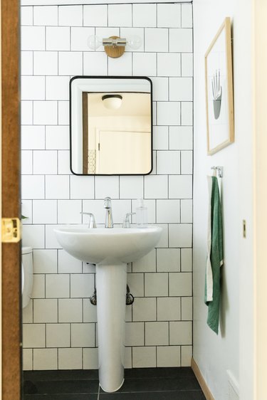 bathroom, large white subway tile wall, white pedestal sink with silver faucet, rectangular mirror with black trim, green hanging towel