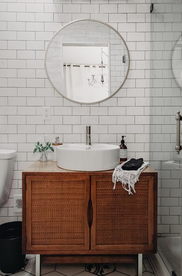 wood vanity with white vessel sink, round mirror with silver trim, white subway tile wall, glass shower door