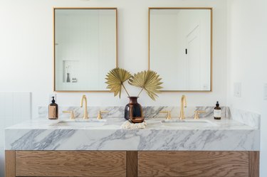 wood bathroom vanity with marble counters, double sinks with gold fixtures, two rectangular mirrors with gold trim