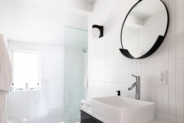 white vessel sink with silver faucet, round mirror with black trim, shower with glass door, white vertical rectangular tile